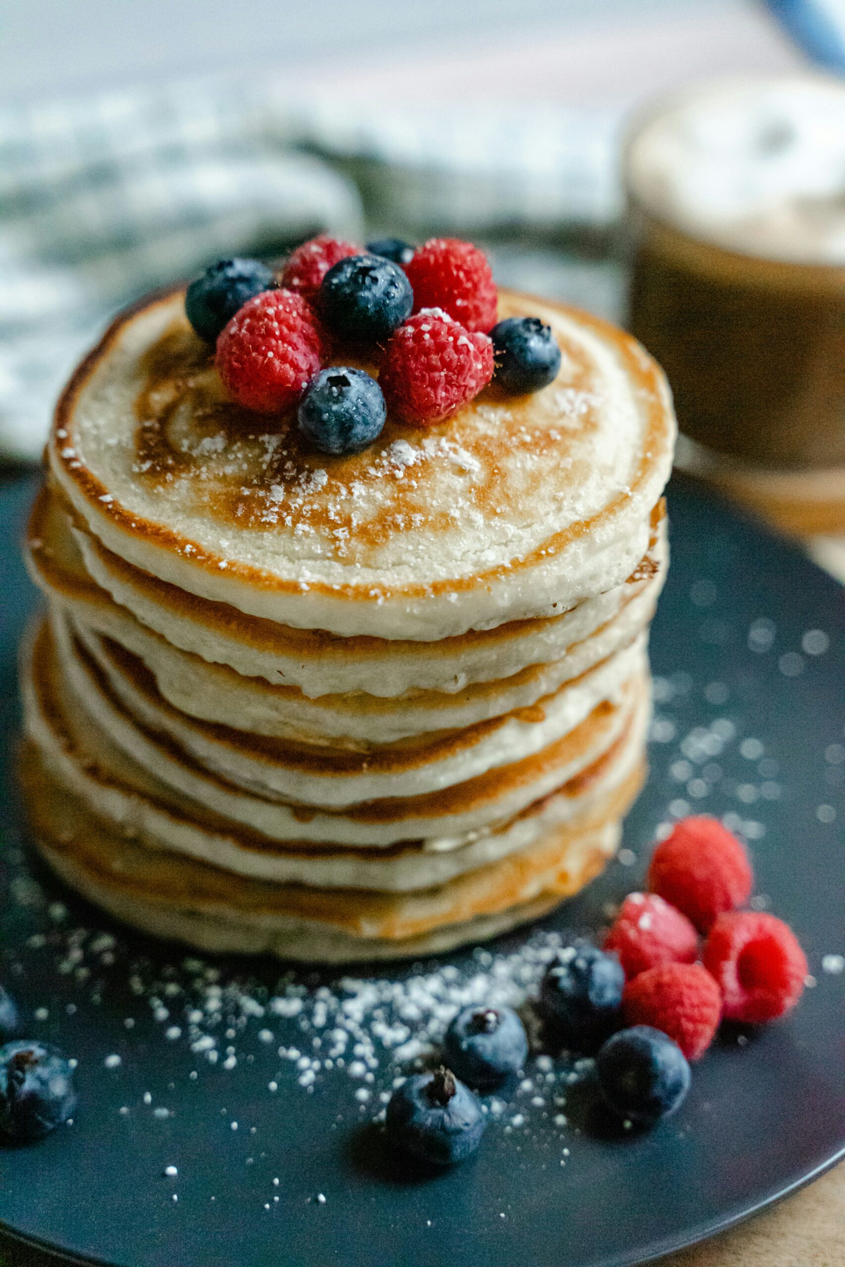 Ein Stapel Pancakes mit Beeren und Puderzucker auf einem dunklen Teller.